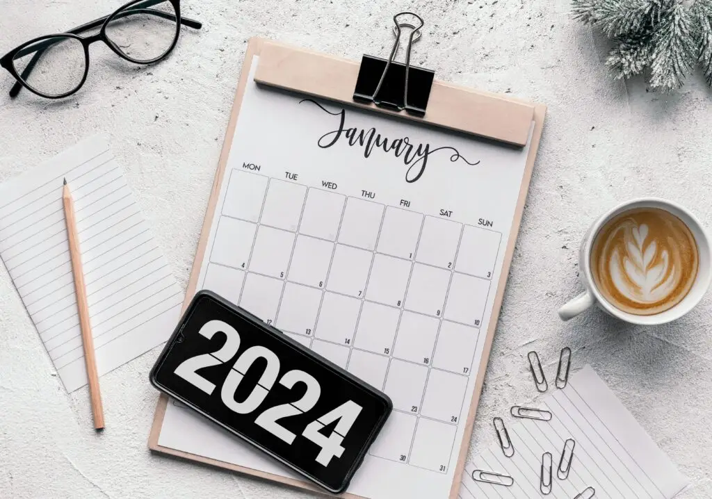 January 2024 calendar on clipboard with glasses, coffee cup, and office supplies around it.