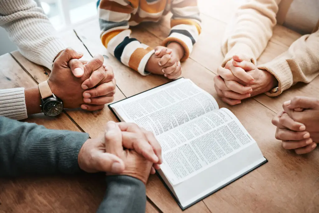 Hands of family folded in prayer around open bible on table