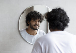 Indian guy looking at his reflection in the mirror, standing in modern bathroom at home.
