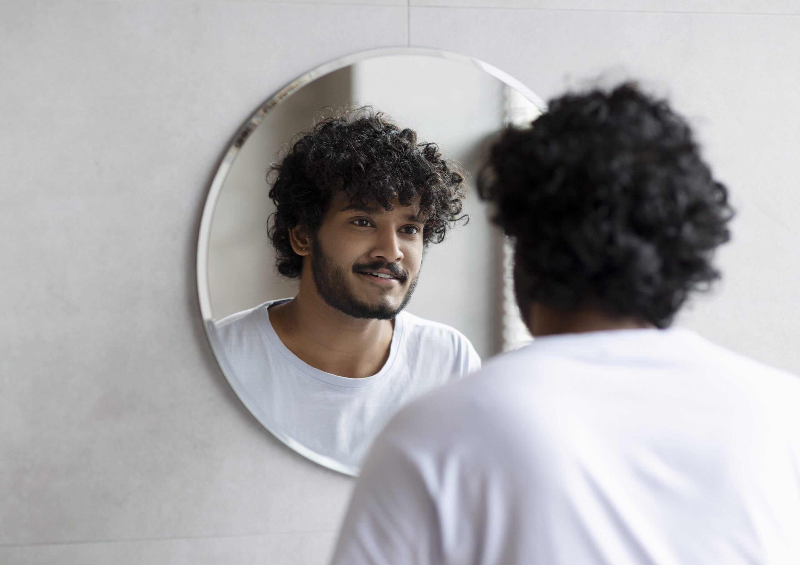 Indian guy looking at his reflection in the mirror, standing in modern bathroom at home.