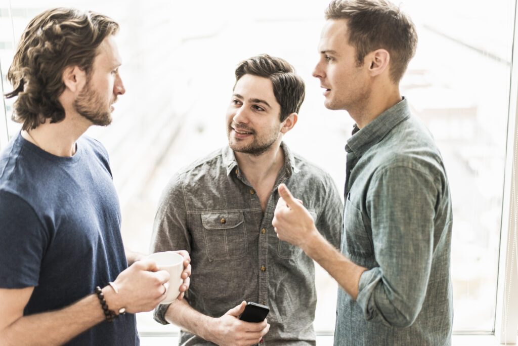 Three men standing talking, one with a cup of coffee, one with a smartphone.